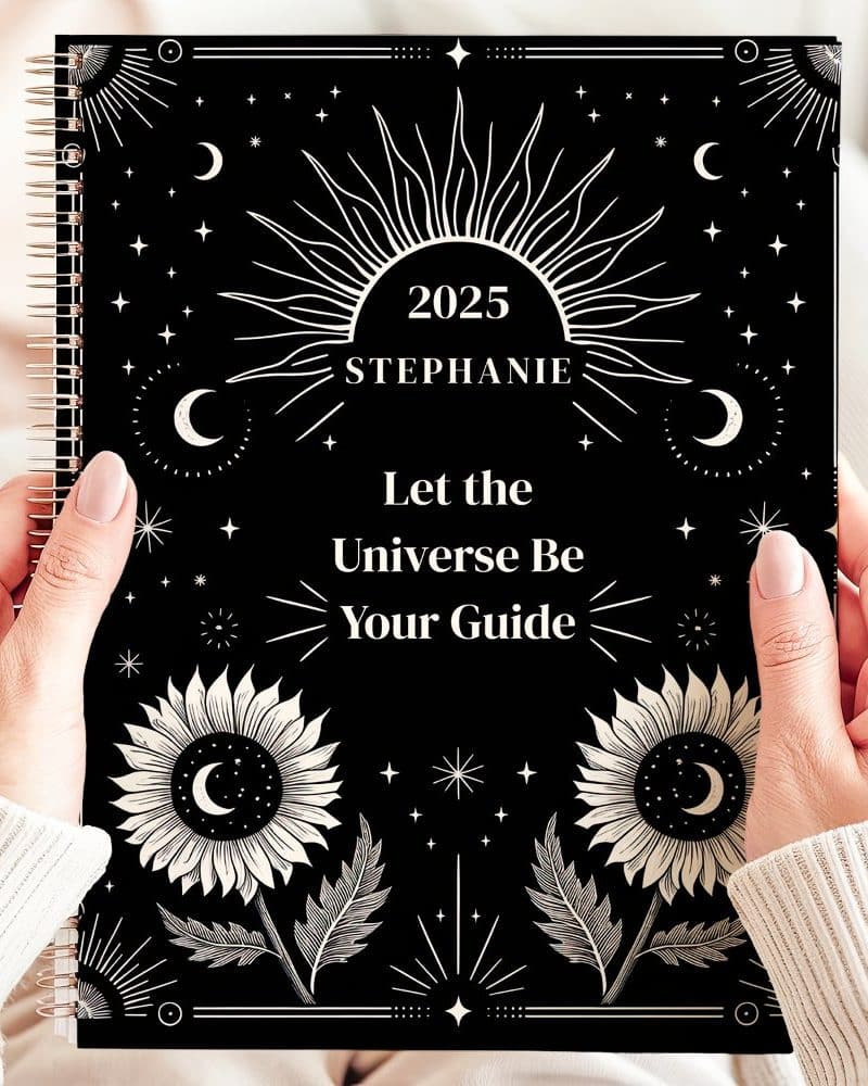 Two hands hold a 2025 planner with a black cover. The cover features a celestial design with suns, moons, stars, and sunflowers in cream. Text reads Let the Universe Be Your Guide. The name Stephanie is also visible.