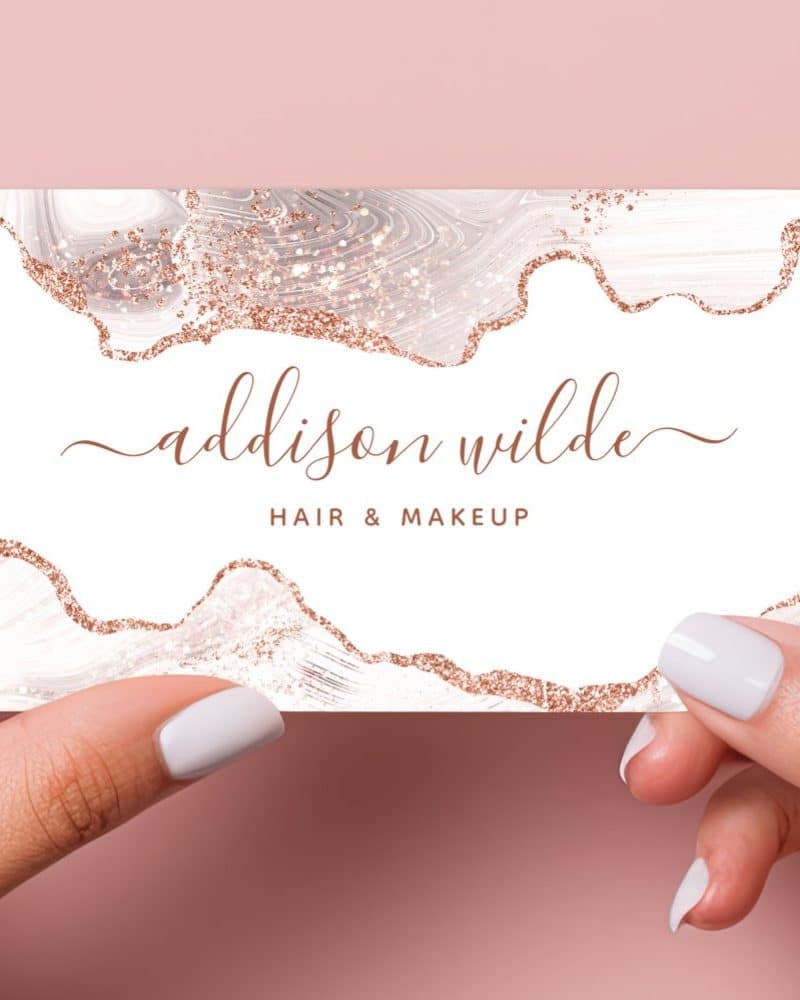 Two hands hold up a business card featuring a rose gold glitter and marble design. The name Addison Wilde, Hair & Makeup is displayed on the card.