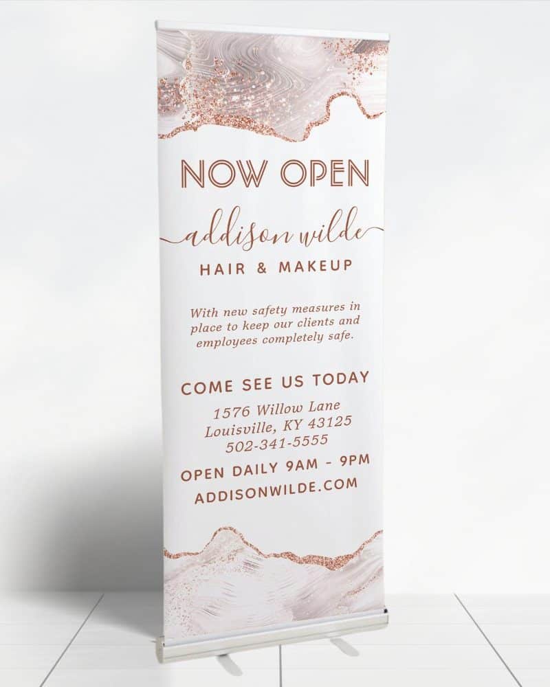 A retractable banner announcing the grand opening of Addison Wilde Hair & Makeup. The banner features a rose gold and white marble design.