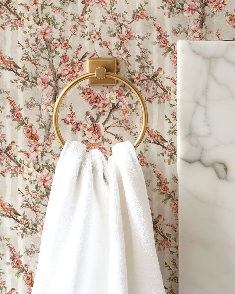 White towel hanging on a gold towel ring against a floral wallpaper.
