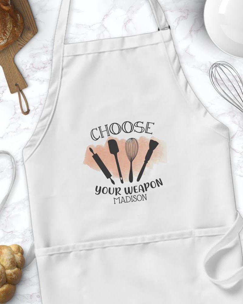 White apron with the text Choose Your Weapon and the name Madison, along with a design of baking utensils.