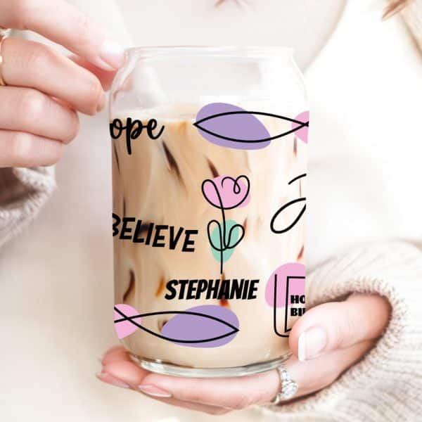 Two Hands Hold A Glass Can-Shaped Iced Coffee Cup With The Name Stephanie And The Words Hope And Believe Printed On It. Pastel Shapes And A Simple Flower Design Decorate The Cup.