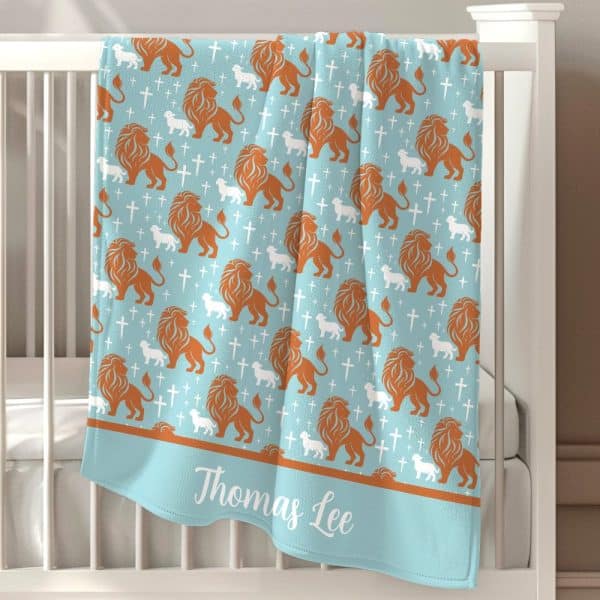 Light Blue Baby Blanket With A Repeating Pattern Of Orange Lions And White Lambs, And White Crosses. The Name Thomas Lee Is Embroidered On The Bottom.