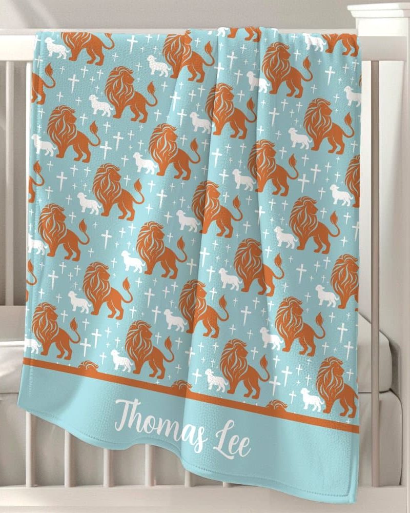 Light blue baby blanket with a repeating pattern of orange lions and white lambs, and white crosses. The name Thomas Lee is embroidered on the bottom.