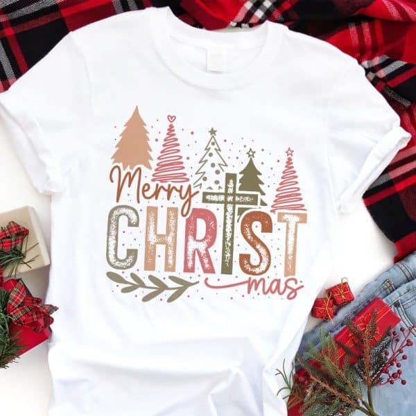 White T-Shirt With A Christmas Design Featuring Stylized Trees, A Cross, And The Text Merry Christmas In A Distressed Font.