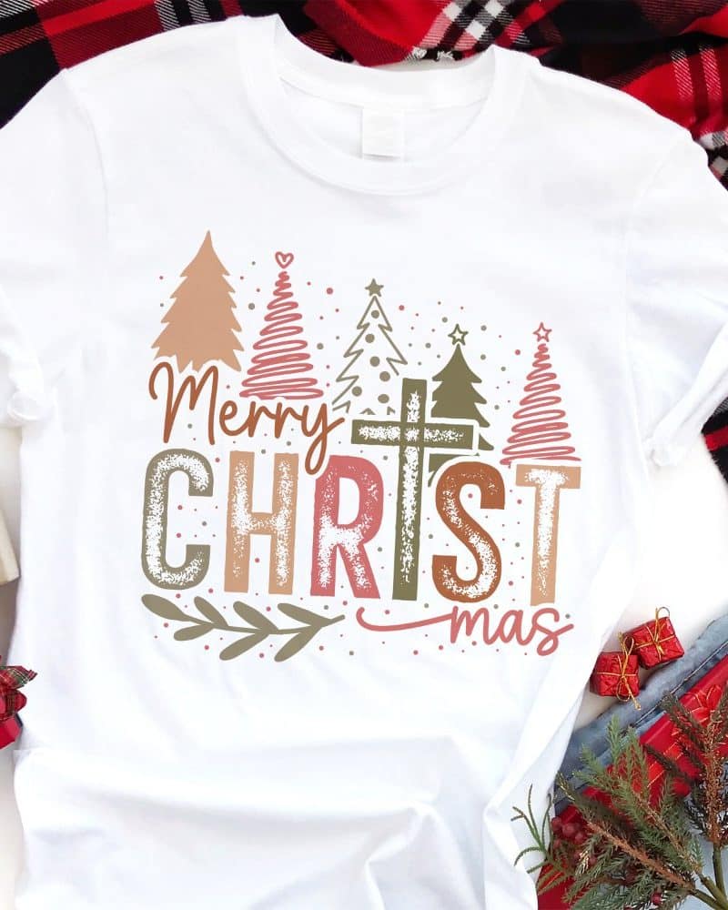 White t-shirt with a Christmas design featuring stylized trees, a cross, and the text Merry Christmas in a distressed font.