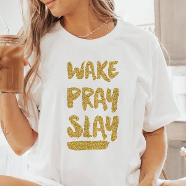 Woman Wearing A White T-Shirt With Gold Glitter Lettering That Reads Wake Pray Slay.