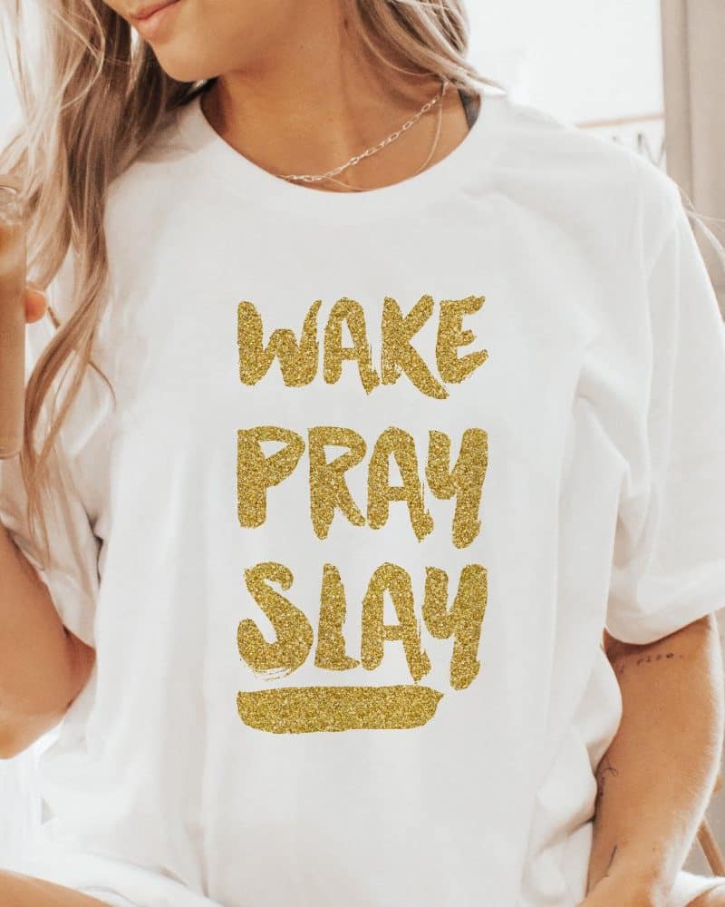 Woman wearing a white t-shirt with gold glitter lettering that reads Wake Pray Slay.