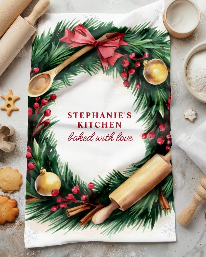 Christmas-themed kitchen tea towel featuring a wreath design with baking utensils and the text Stephanie's Kitchen baked with love.