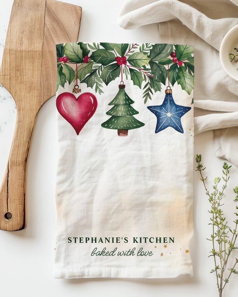 Christmas tea towel with a holly border and hanging ornaments: a heart, tree, and star. The text, Stephanie's Kitchen, baked with love, is at the bottom.