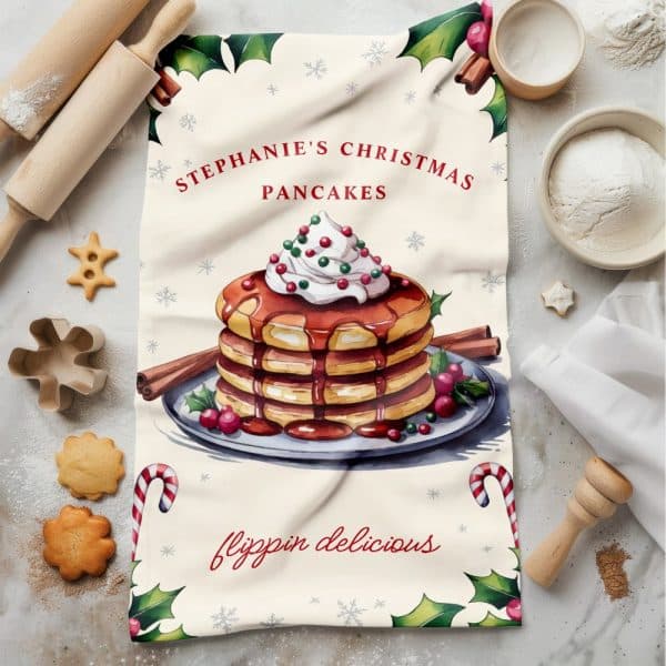 Christmas-Themed Tea Towel Featuring A Watercolor Illustration Of A Stack Of Pancakes Topped With Whipped Cream And Syrup.