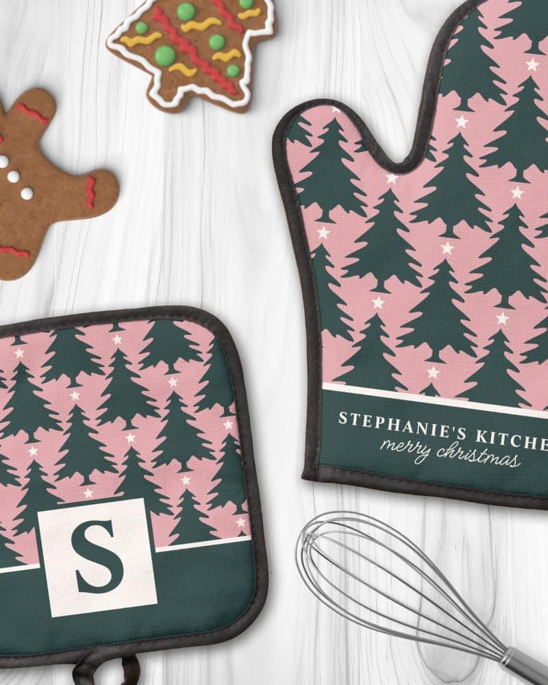 A pair of Christmas-themed oven mitts and a pot holder. The pink and dark green mitts and holder feature a repeating pattern of Christmas trees and stars. One mitt is personalized with a name and Merry Christmas.