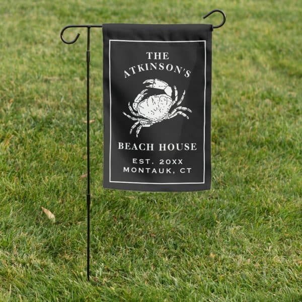 Black Garden Flag With A White Crab Illustration, The Atkinsons Beach House Text, And Establishment Year And Location.
