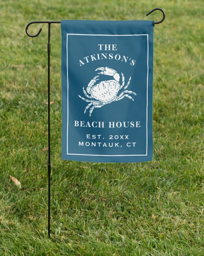 A dark blue garden flag with a white crab illustration, text reading The Atkinsons Beach House Est. 20XX Montauk, CT.