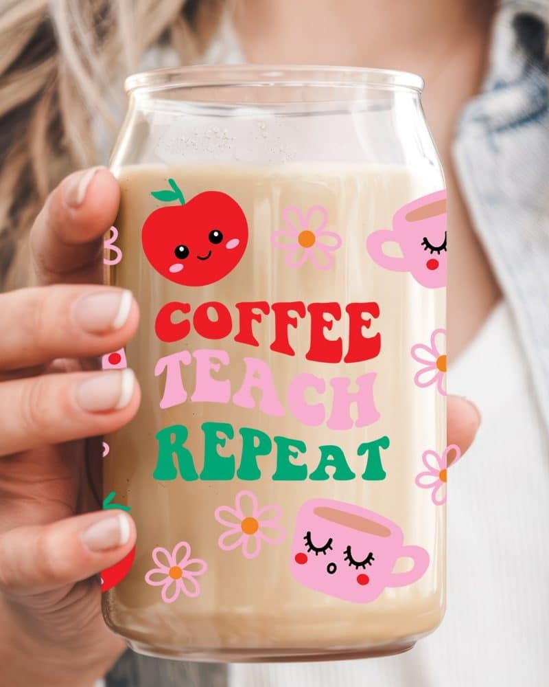 Person holding a glass with coffee and the words Coffee Teach Repeat. The glass is decorated with cartoon apples and coffee cups.