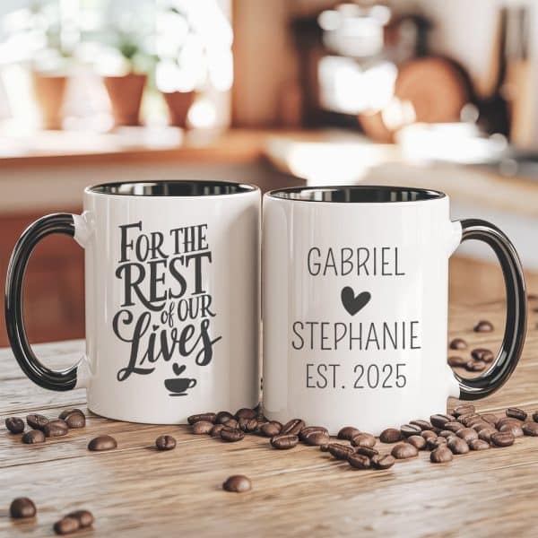 Two Personalized Coffee Mugs With Black Handles And Rims Sit On A Wood Surface Surrounded By Coffee Beans. One Mug Says For The Rest Of Our Lives, The Other Says Gabriel And Stephanie Est. 2025.