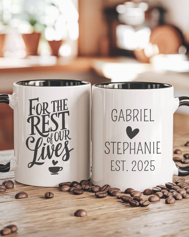 Two personalized coffee mugs with black handles and rims sit on a wood surface surrounded by coffee beans. One mug says For the Rest of Our Lives, the other says Gabriel and Stephanie Est. 2025.
