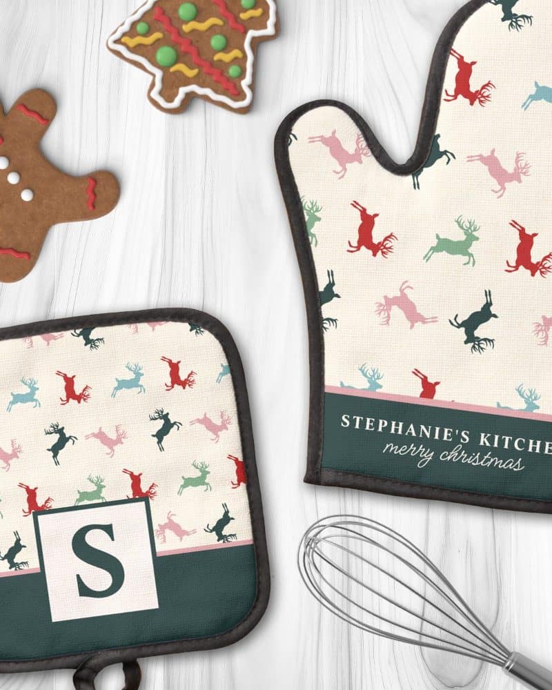 A personalized oven mitt and pot holder with a cream background and a repeating pattern of colorful reindeer. The oven mitt is labeled "Stephanie's Kitchen Merry Christmas". The pot holder has a monogrammed "S".