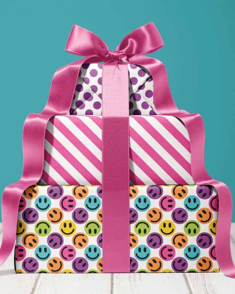 A stack of three presents tied with a large pink ribbon. The top present has purple polka dots, the middle has pink stripes, and the bottom has colorful smiley faces.