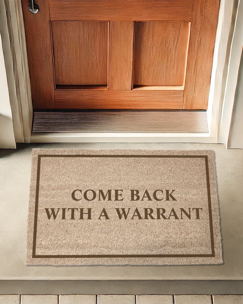 Beige doormat with brown border and text reading Come Back With A Warrant in front of a wooden door.
