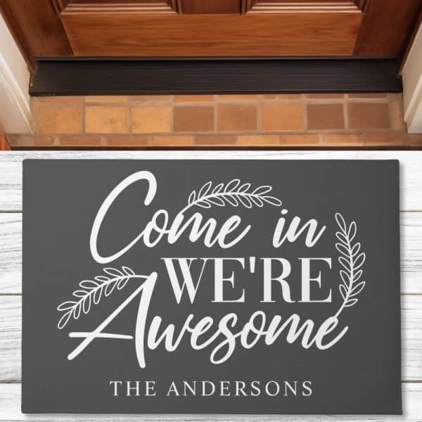 Dark Gray Doormat With White Lettering That Says Come In We'Re Awesome, And The Name The Andersons Below.