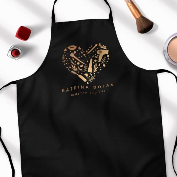 Black Apron With Gold Heart-Shaped Logo Featuring Beauty Tools And The Name Katrina Dolan, Master Stylist.