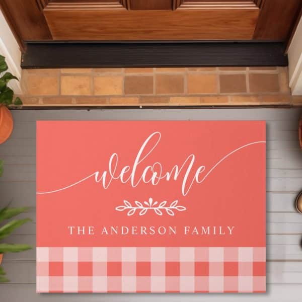 Coral Doormat With White Script That Says Welcome, A Small Floral Design, And The Anderson Family. The Bottom Features A Coral And White Gingham Pattern.