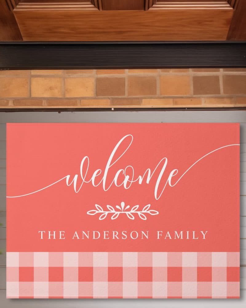 Coral doormat with white script that says Welcome, a small floral design, and The Anderson Family. The bottom features a coral and white gingham pattern.