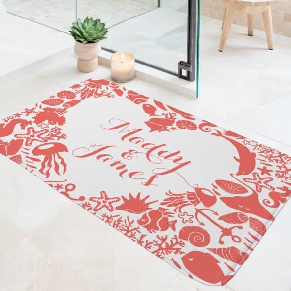 A Personalized Bath Mat With The Names Mady And James In Coral-Colored Script On A White Background. The Names Are Surrounded By A Border Of Coral-Colored Sea Life Silhouettes.