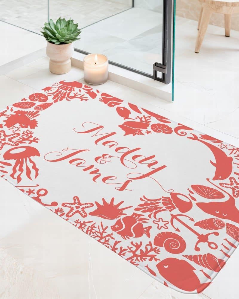 A personalized bath mat with the names Mady and James in coral-colored script on a white background. The names are surrounded by a border of coral-colored sea life silhouettes.