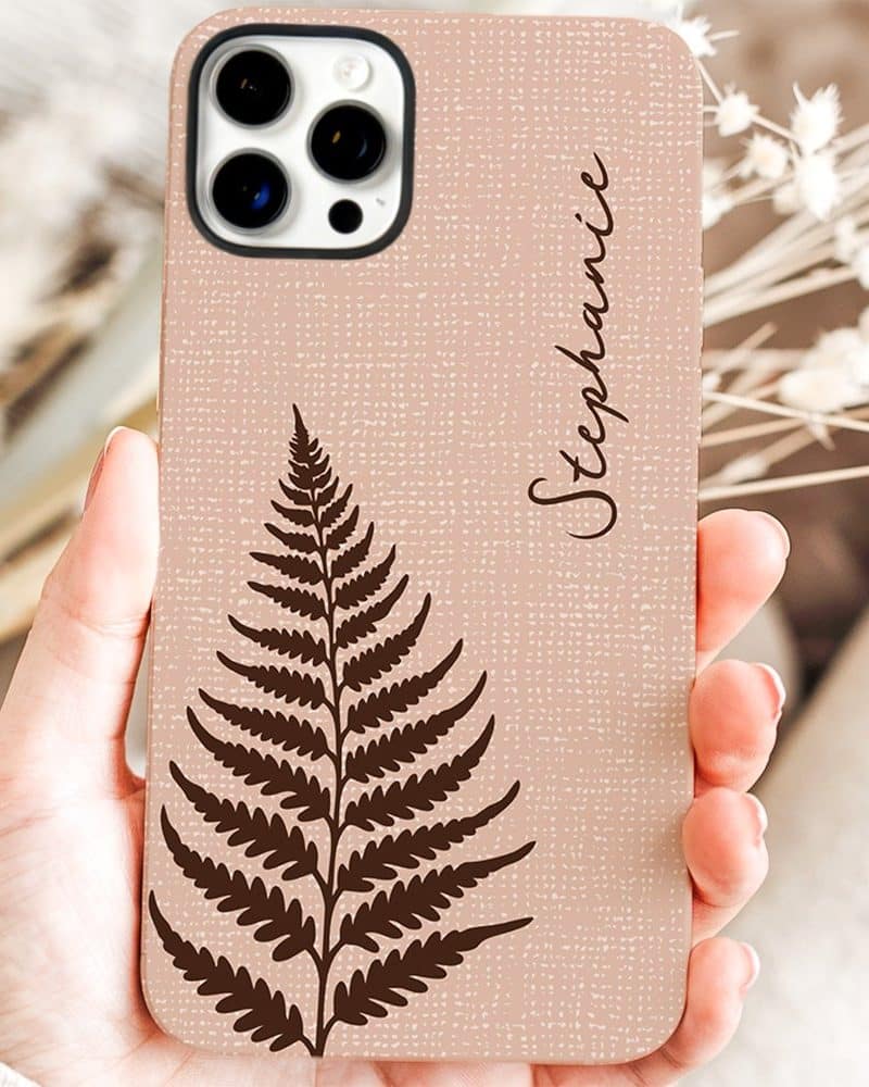 A hand holds a blush-pink phone case featuring a dark brown fern and the name Stephanie.
