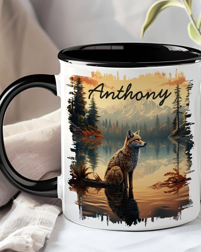 A black and white mug with the name Anthony and a fox sitting by a lake in a mountain landscape.
