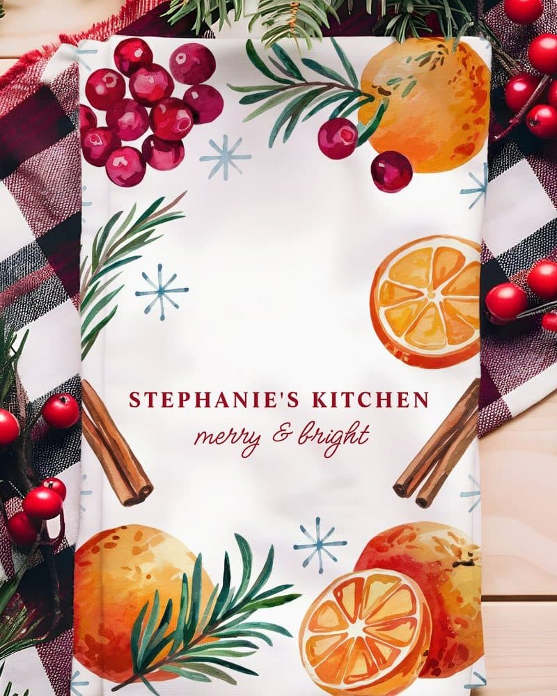 Christmas kitchen tea towel with oranges, cranberries, rosemary, and cinnamon sticks. The text "Stephanie's Kitchen merry & bright" is also on the towel.