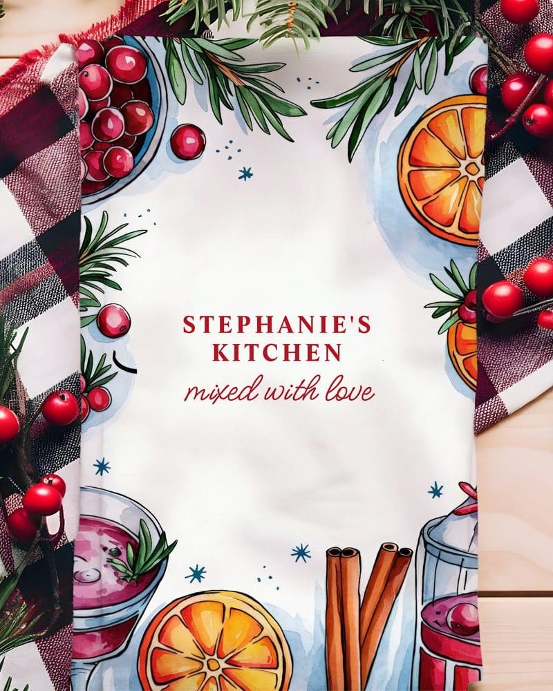 A kitchen tea towel featuring watercolor illustrations of cranberries, oranges, cinnamon sticks, and warm drinks, with the text Stephanie's Kitchen, mixed with love.