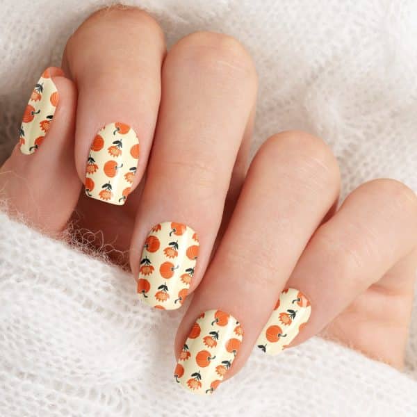 A Hand Displaying Fingernails With A Fall-Themed Design Of Small Orange Pumpkins And Flowers On A Cream Background.