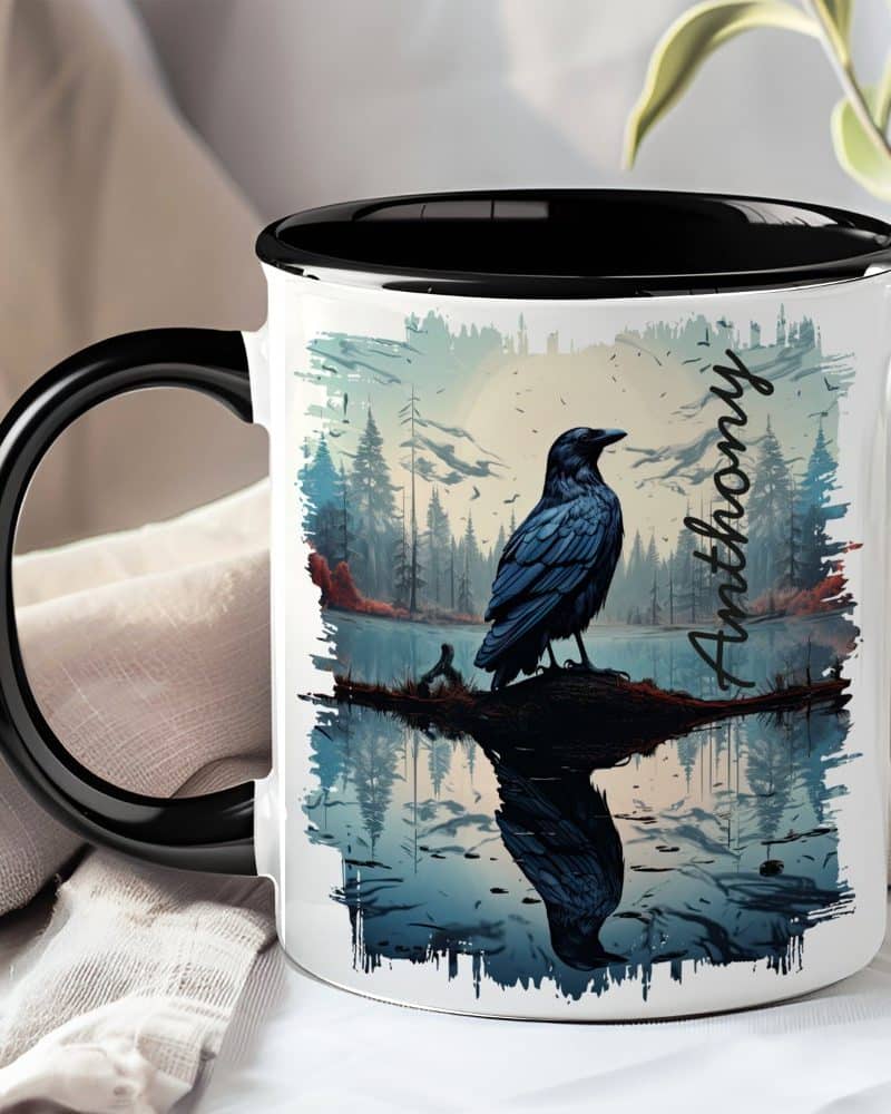 A black mug with a black handle features a raven perched on a branch over water, reflected in the still surface. The name Anthony is printed on the mug.