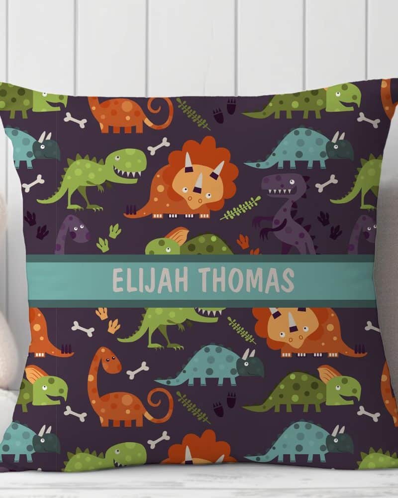 Personalized square pillow with a repeating pattern of colorful cartoon dinosaurs on a dark purple background. The name Elijah Thomas is printed across the center.