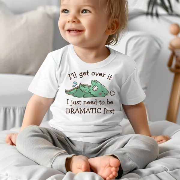 A Toddler Sits On A Bed Wearing A White Shirt Featuring A Cartoon Illustration Of A Small, Injured Dragon And The Text, Ill Get Over It I Just Need To Be Dramatic First.
