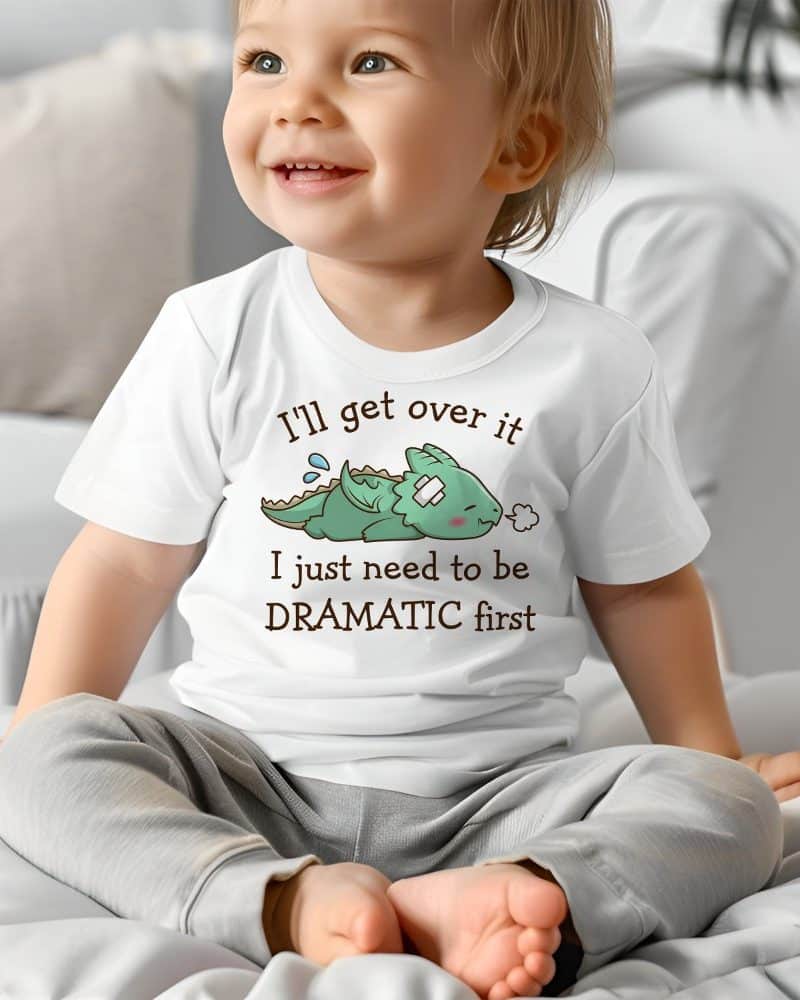 A toddler sits on a bed wearing a white shirt featuring a cartoon illustration of a small, injured dragon and the text, Ill get over it I just need to be DRAMATIC first.