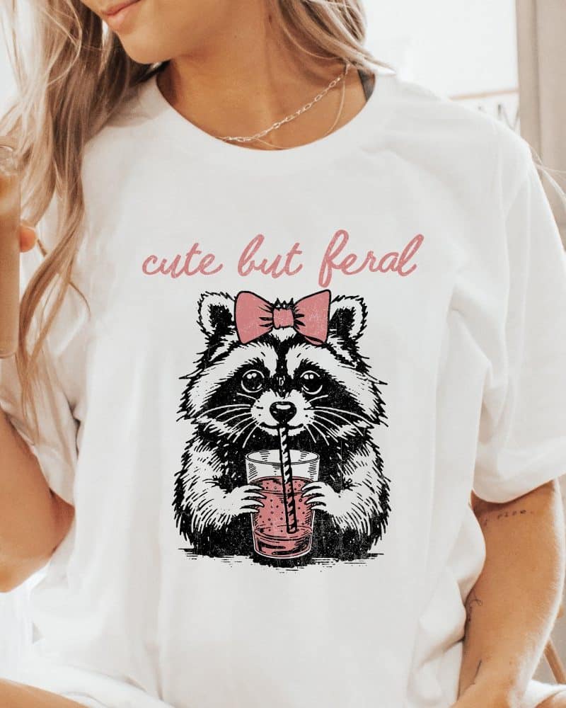 White t-shirt featuring a drawing of a raccoon wearing a bow and drinking from a glass with a straw. Text above reads cute but feral.