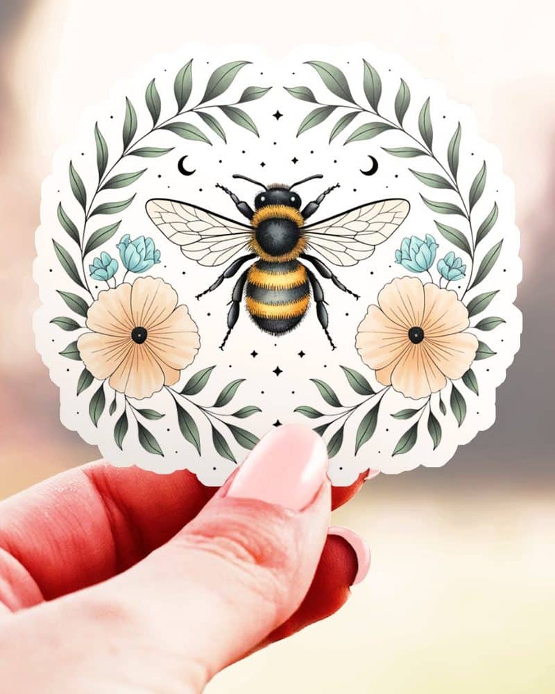 A hand holds a circular sticker featuring a bumble bee centered in a floral wreath.