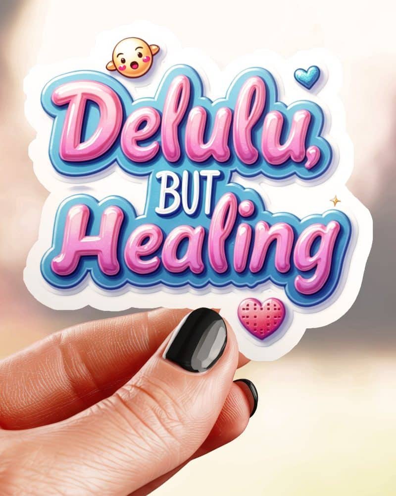 A hand holds a sticker that says Delulu, But Healing in stylized pink and blue lettering.