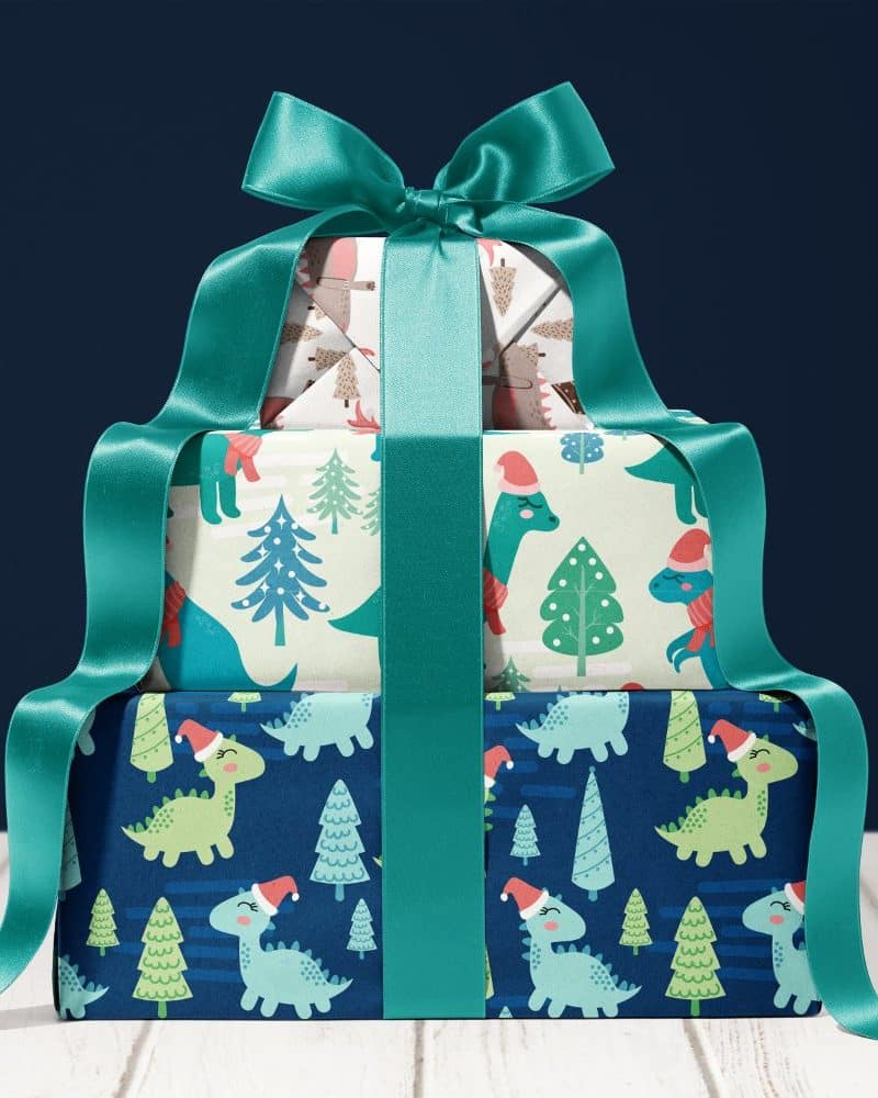 Three Christmas presents stacked, wrapped in dinosaur and Christmas tree patterned paper, tied with a teal ribbon.