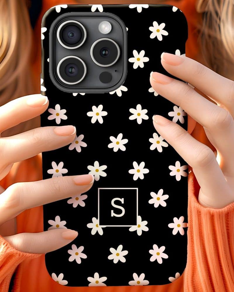 A woman's hands hold a black phone case covered in small white daisies and a white square with the letter S in the center.