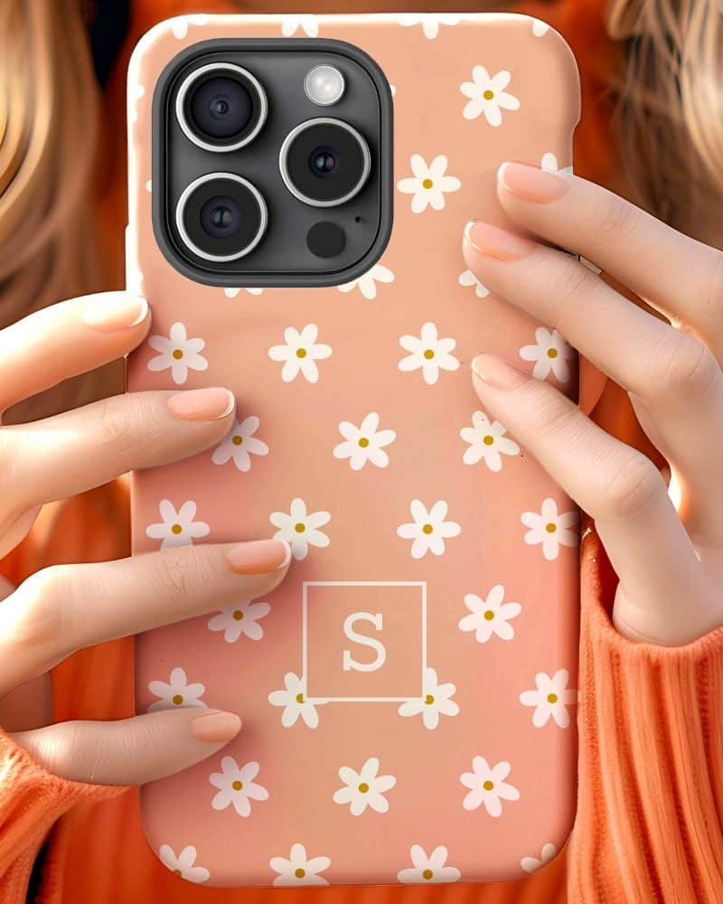 Close-up of hands holding a peach-colored phone case with white daisies and a monogram.