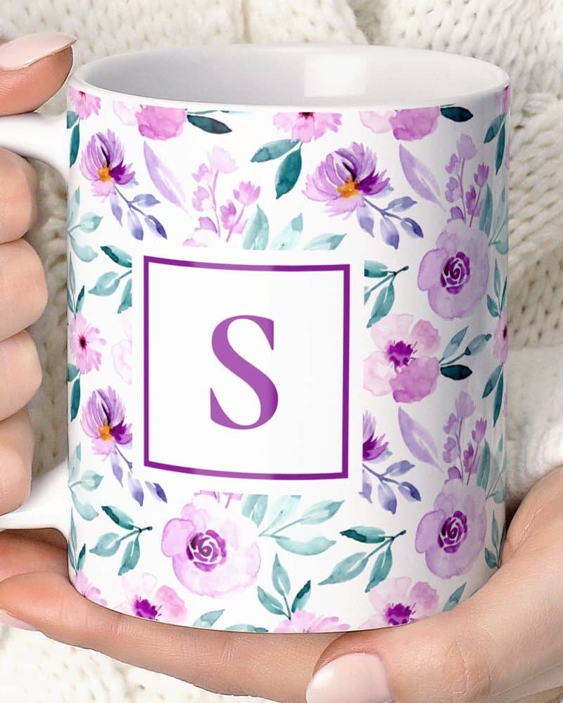 A white mug with a purple floral pattern and a purple letter S in a box.