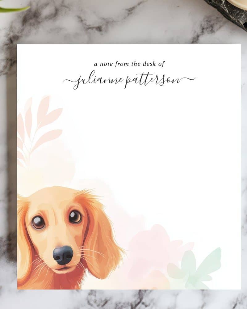 A tan longhaired dachshund's head and shoulders peek from behind pastel flowers on a personalized notepad.