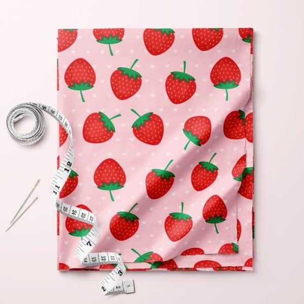Pink Fabric With A Repeating Pattern Of Red Strawberries And Small White Hearts. A Tape Measure And Needles Are Beside It.
