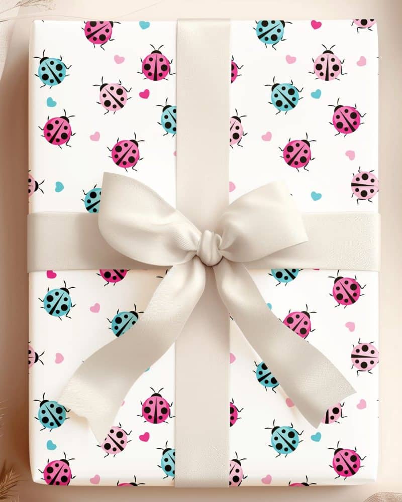 Gift wrapped in white paper with a repeating pattern of pink, blue, and black ladybugs and small hearts. A beige ribbon is tied in a bow on top.