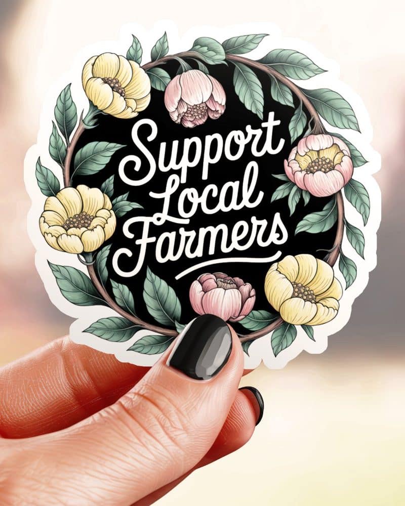 A hand holds a round sticker with a floral wreath surrounding the text Support Local Farmers.
